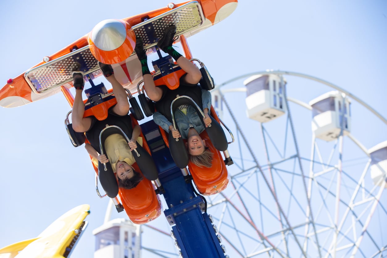 Two people are upside down on an orange amusement park ride, smiling and holding on. The background shows a Ferris wheel against a clear blue sky.