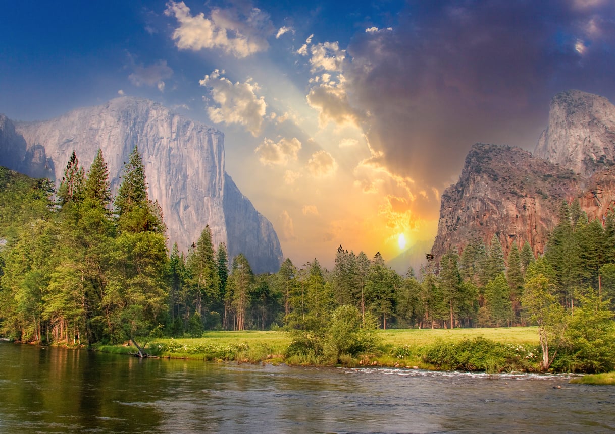 Sunset over a river in Yosemite Valley, with pine trees, green meadow, and dramatic cliffs under a colorful, partly cloudy sky.