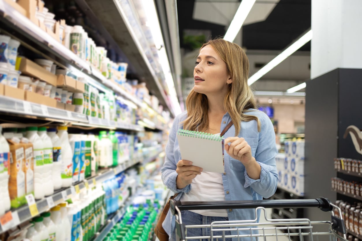A woman holding a notepad stands in a grocery store aisle, looking at shelves of dairy products while pushing a shopping cart.