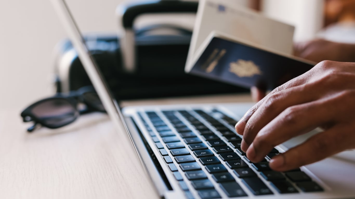 Hands typing on a laptop keyboard with a passport and boarding pass nearby; sunglasses and a suitcase are also visible in the background, suggesting travel preparations before booking a flight.