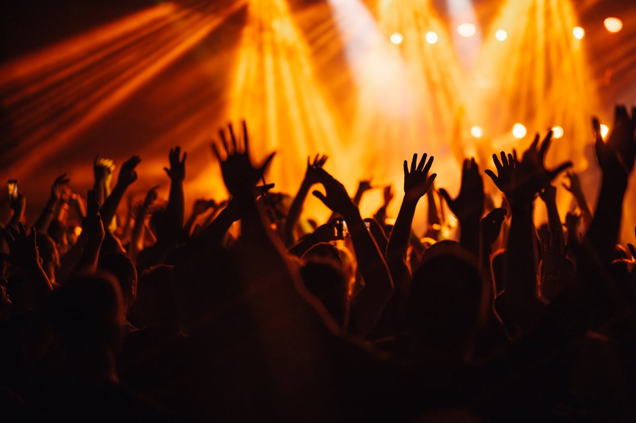 A crowd of people at a concert with their hands raised, illuminated by orange stage lights in the background.
