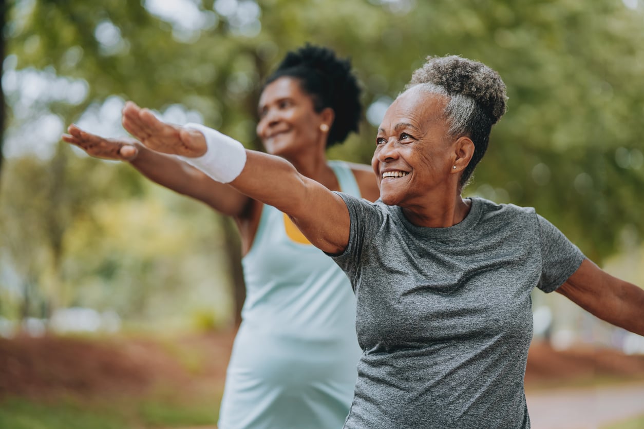 Two older women smiling and practicing yoga outdoors, standing with arms extended and looking focused. Trees and greenery are visible in the background, reflecting how Women Are Changing Their Spending Behaviors by investing in health and well-being activities.
