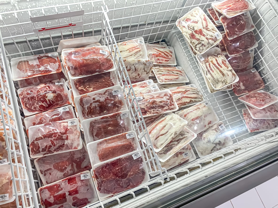 Packages of assorted raw meats, including beef and pork, are neatly arranged in a supermarket freezer display, all wrapped in clear plastic and placed on white trays.
