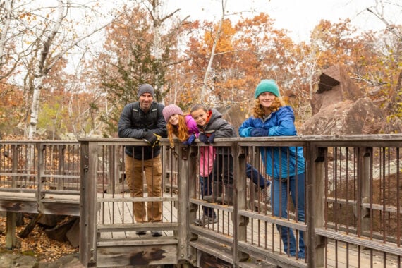 Four people in warm jackets and hats lean on a wooden railing of a bridge in a forest with autumn leaves. Smiling and posing for the photo, they look like friends enjoying an outing&mdash;perhaps making memories together as part of one of those memberships worth paying for.