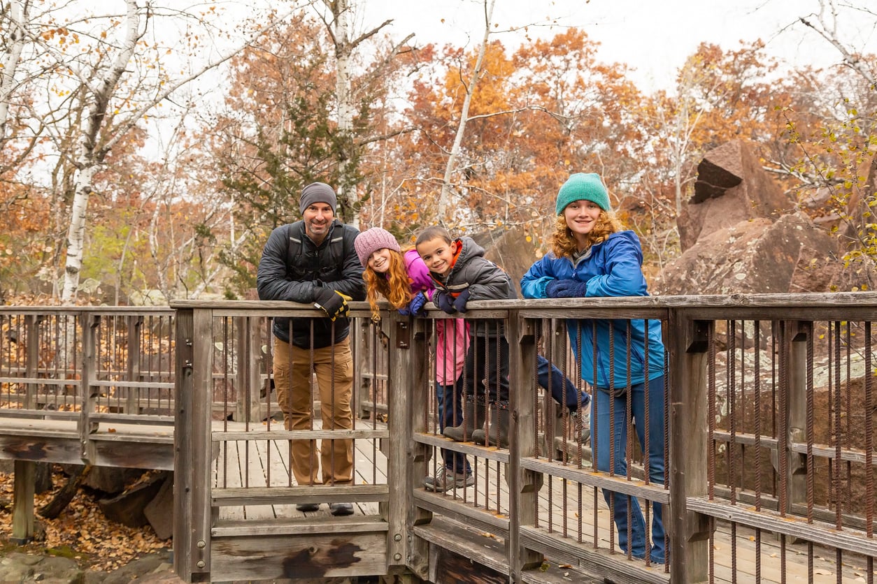 Four people in warm jackets and hats lean on a wooden railing of a bridge in a forest with autumn leaves. Smiling and posing for the photo, they look like friends enjoying an outing—perhaps making memories together as part of one of those memberships worth paying for.