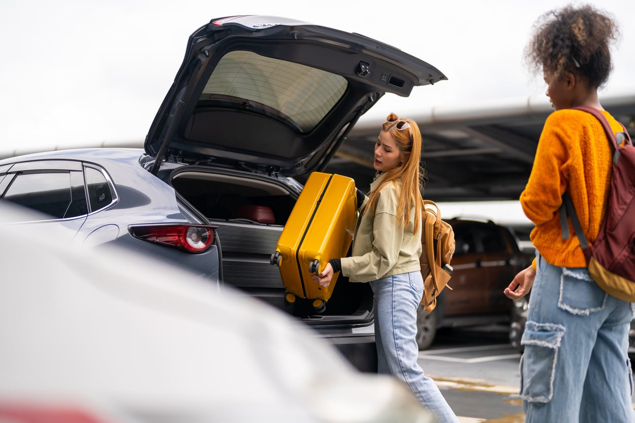 Two people stand by an open car trunk. One person places a yellow suitcase inside, while the other watches. Both are wearing casual clothes and backpacks in a parking lot.