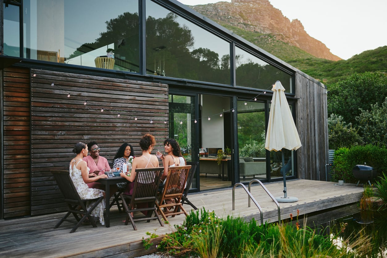 Five women sit at a wooden table outside a modern house, enjoying food and drinks surrounded by stylish outdoor home products. String lights hang overhead, casting a warm glow, while mountains and greenery provide a beautiful backdrop.
