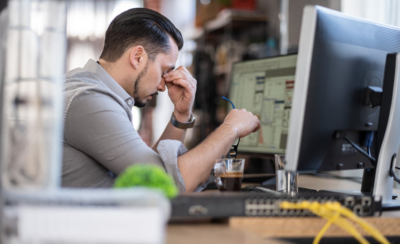 A man sits at a desk in front of a computer, holding his glasses and rubbing his forehead, appearing stressed or tired. There is a coffee cup and computer equipment on the desk.