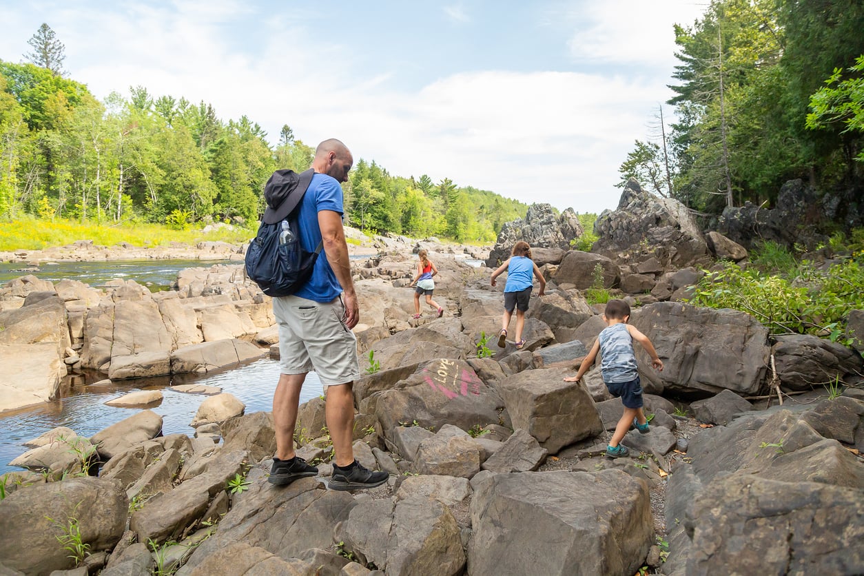 A man and four children climb over large rocks by a river surrounded by trees on a sunny day, enjoying quality time together—experiences like these make memberships worth paying for.