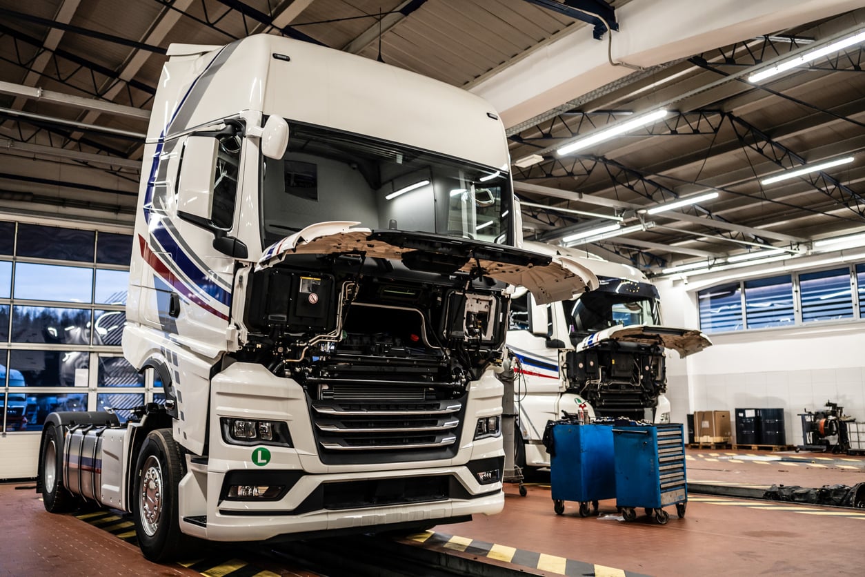 Two large white trucks with open hoods are parked inside a brightly lit garage or service center, where tool carts and various products are positioned nearby for maintenance work.