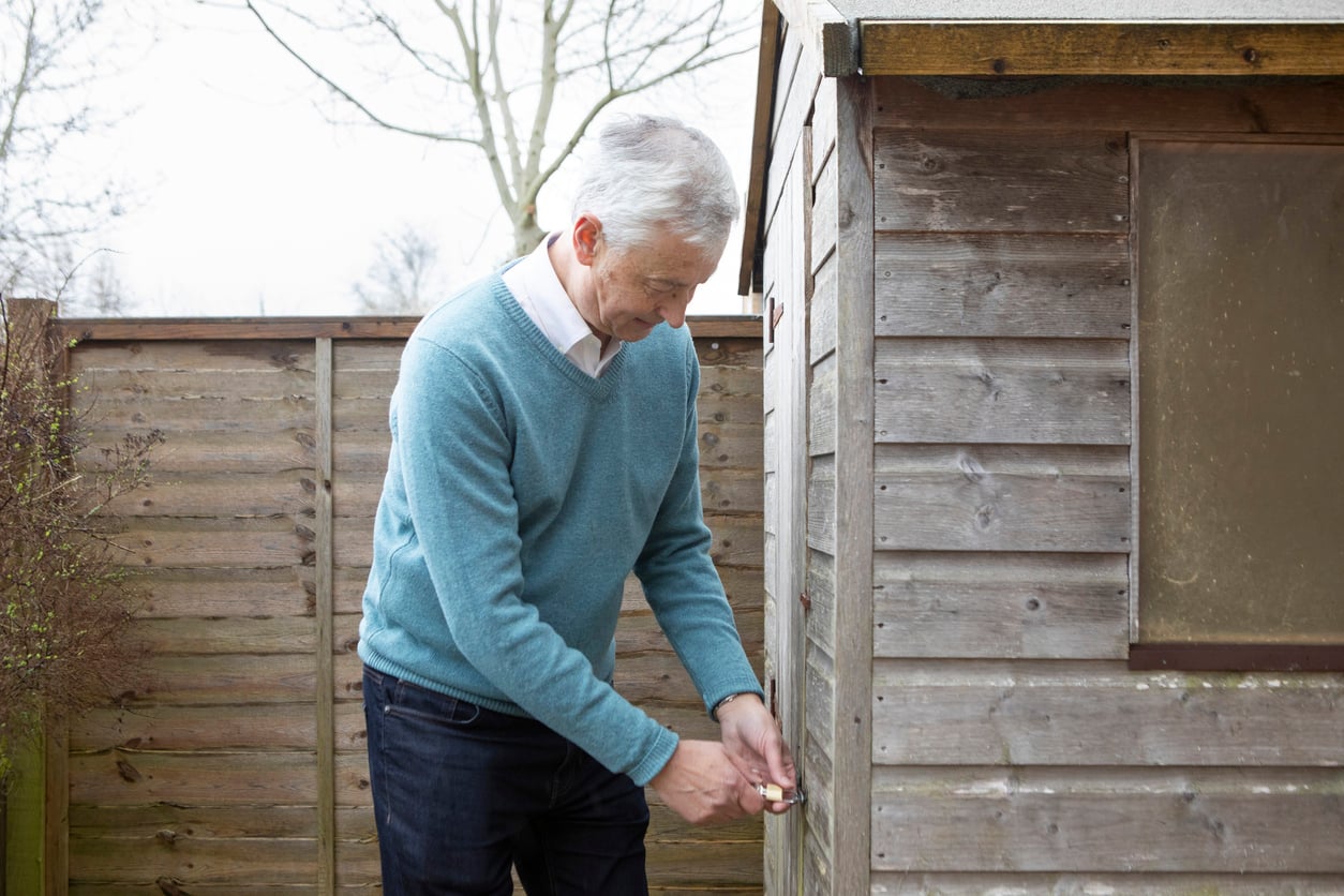 An older man with gray hair, wearing a blue sweater and jeans, unlocks the door of a wooden garden shed—one of many essential outdoor home products—next to a wooden fence on a cloudy day.