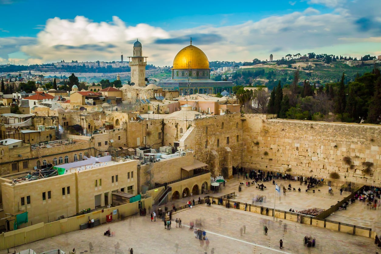 Wide view of Jerusalem’s Old City showing the Western Wall in the foreground, people gathered nearby, and the golden Dome of the Rock with a cloudy blue sky in the background—a stunning scene that highlights why Jerusalem is one of the world’s most captivating travel destinations.