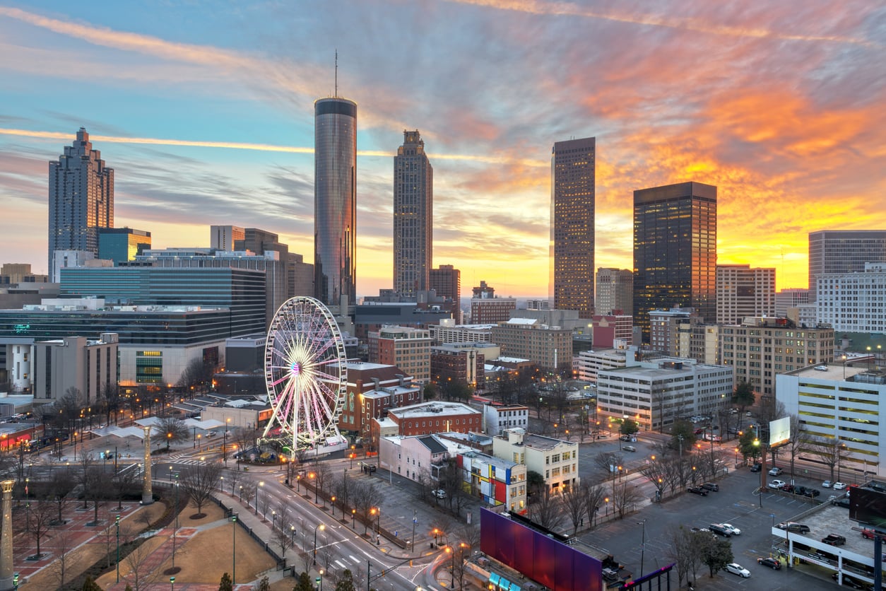 Downtown Atlanta skyline at sunset with tall buildings, a Ferris wheel, and colorful clouds in the sky. The city lights are starting to turn on, showcasing why Atlanta stands out among the big cities in America, and the scene includes a mix of modern and historic architecture.
