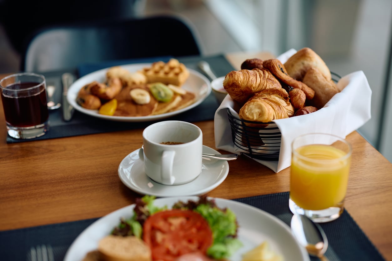 A breakfast table reflecting recent hotel changes, featuring a cup of coffee, orange juice, fresh salad and eggs, waffles with fruit, an extra glass of juice, and a basket of croissants and pastries.