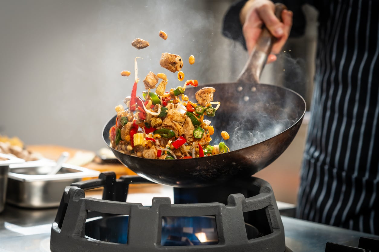 A chef tosses vegetables and meat in a wok over a gas flame, with colorful ingredients mid-air and steam rising from the pan.
