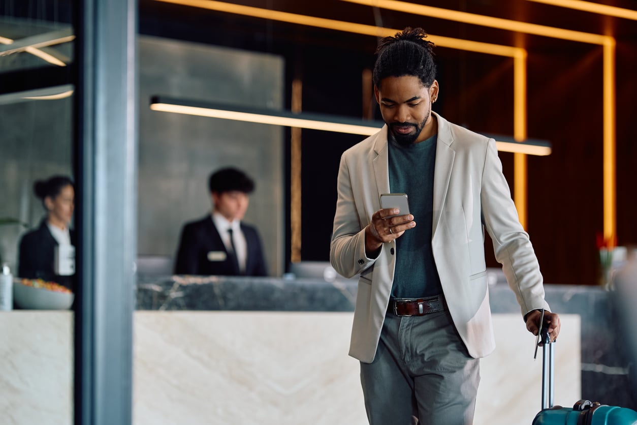 A man in a light suit checks his phone—perhaps reviewing some last-minute hotel booking tips—while pulling a suitcase through a hotel lobby. Two receptionists stand behind the marble front desk in the background.
