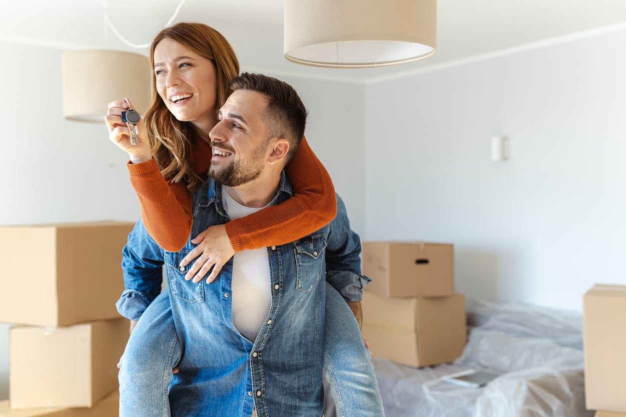 A smiling woman on a man’s back holds up keys in a room with moving boxes, suggesting they’ve just moved into a new home and are celebrating this milestone in line with Baby Boomer beliefs about the importance of home ownership and new beginnings.