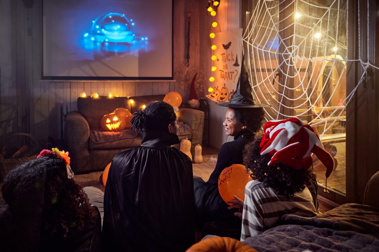 Four kids in Halloween costumes watch a movie in a cozy, decorated living room with pumpkins, candles, a spiderweb on the window, and a “Trick or Treat?” sign. Blue light glows from the screen as they enjoy one of their favorite Halloween activities together.