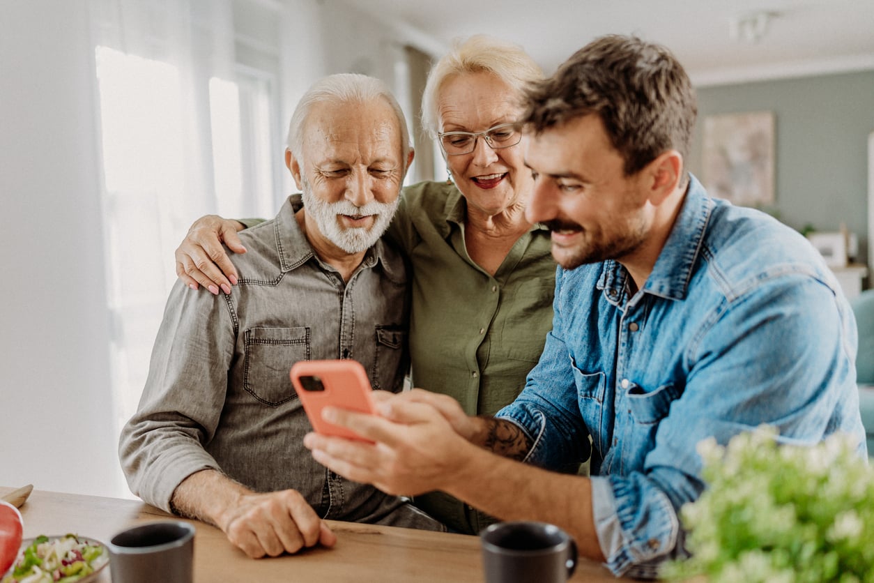 A younger man shows something on a smartphone to an elderly couple at a table. All three are smiling and sitting close together in a bright, cozy room, as they share stories and Baby Boomer beliefs across generations.