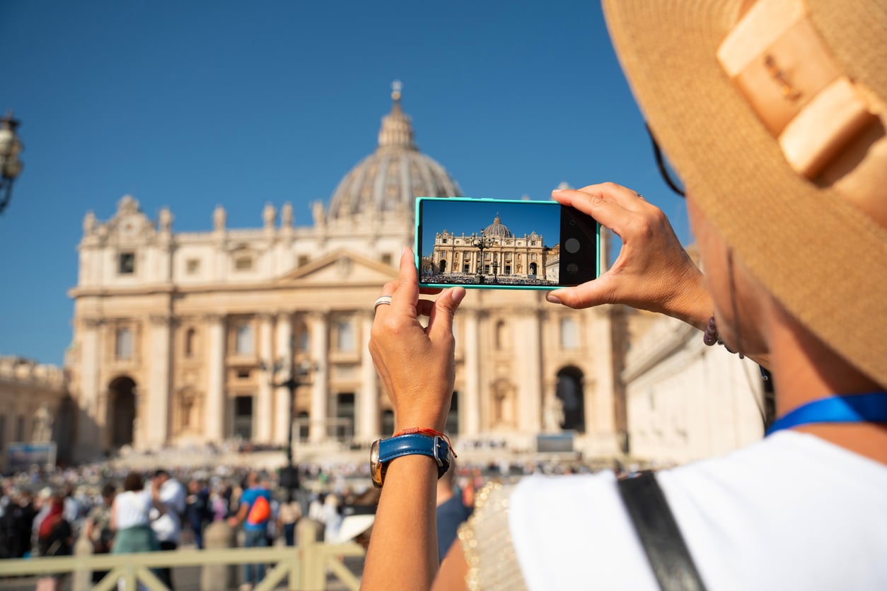A person wearing a hat takes a photo of St. Peter’s Basilica in Vatican City with a smartphone on a sunny day.
