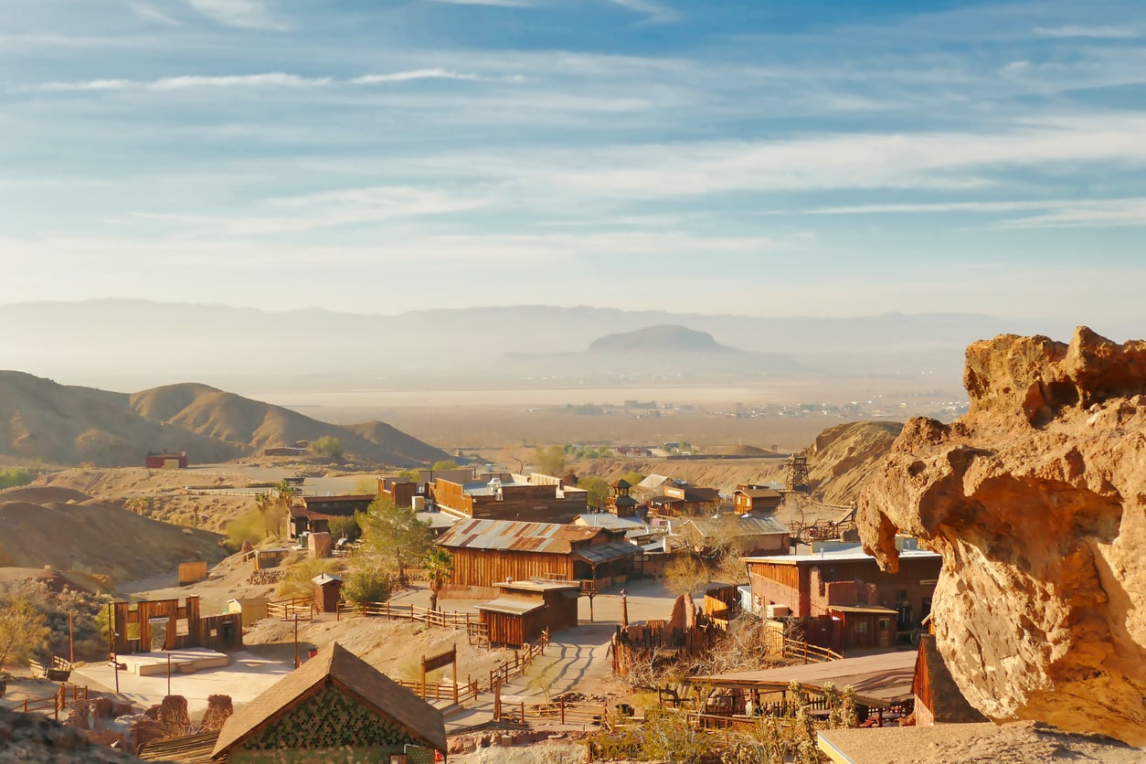 A rustic desert town reminiscent of classic American towns, with wooden buildings and rocky terrain, set against distant mountains under a hazy blue sky.