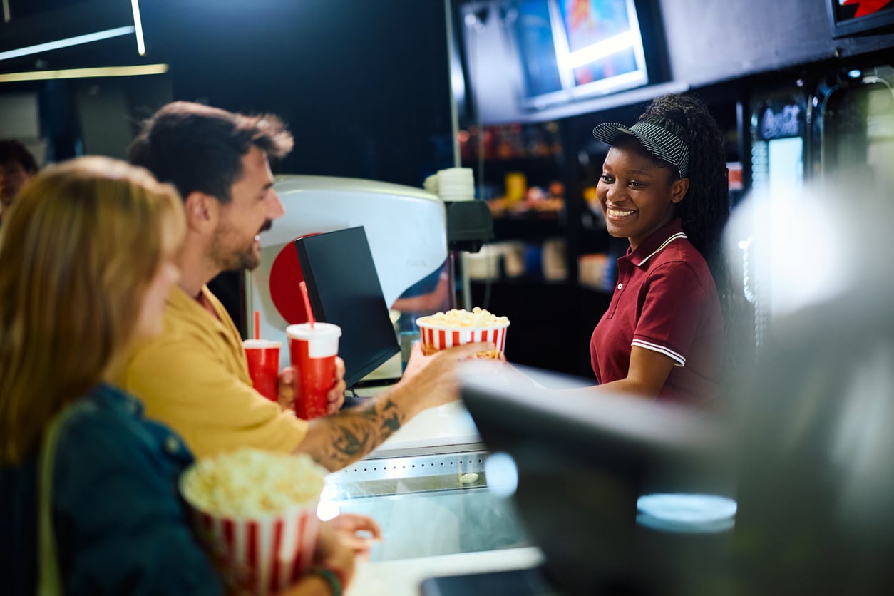 A smiling concession stand worker hands popcorn to a couple at a movie theater counter. They hold drinks and popcorn—classic treats that have endured even as many products people no longer buy have faded away—and the atmosphere is bright and cheerful.