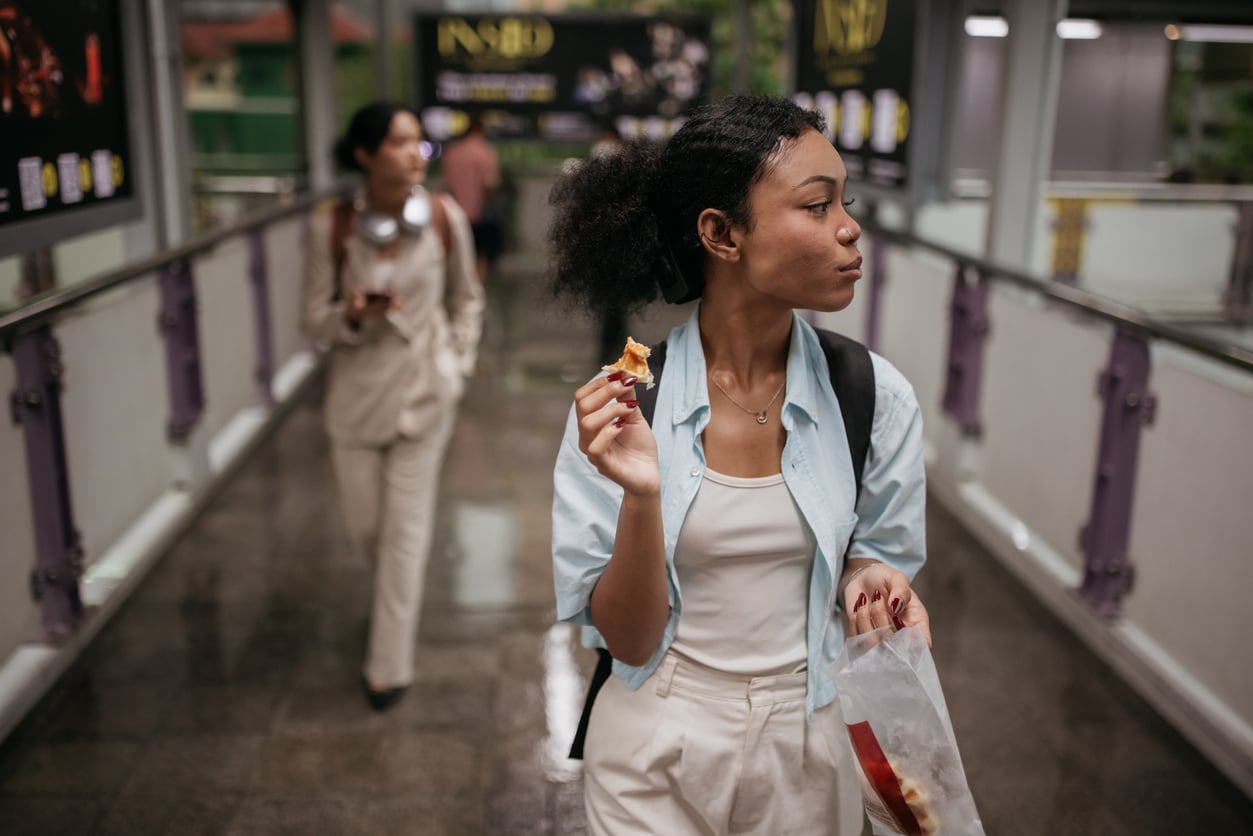 A young woman, possibly reflecting her grocery shopping habits, eats a snack while walking on a covered pedestrian bridge, carrying a backpack and a paper bag. Another person walks behind her, slightly out of focus.