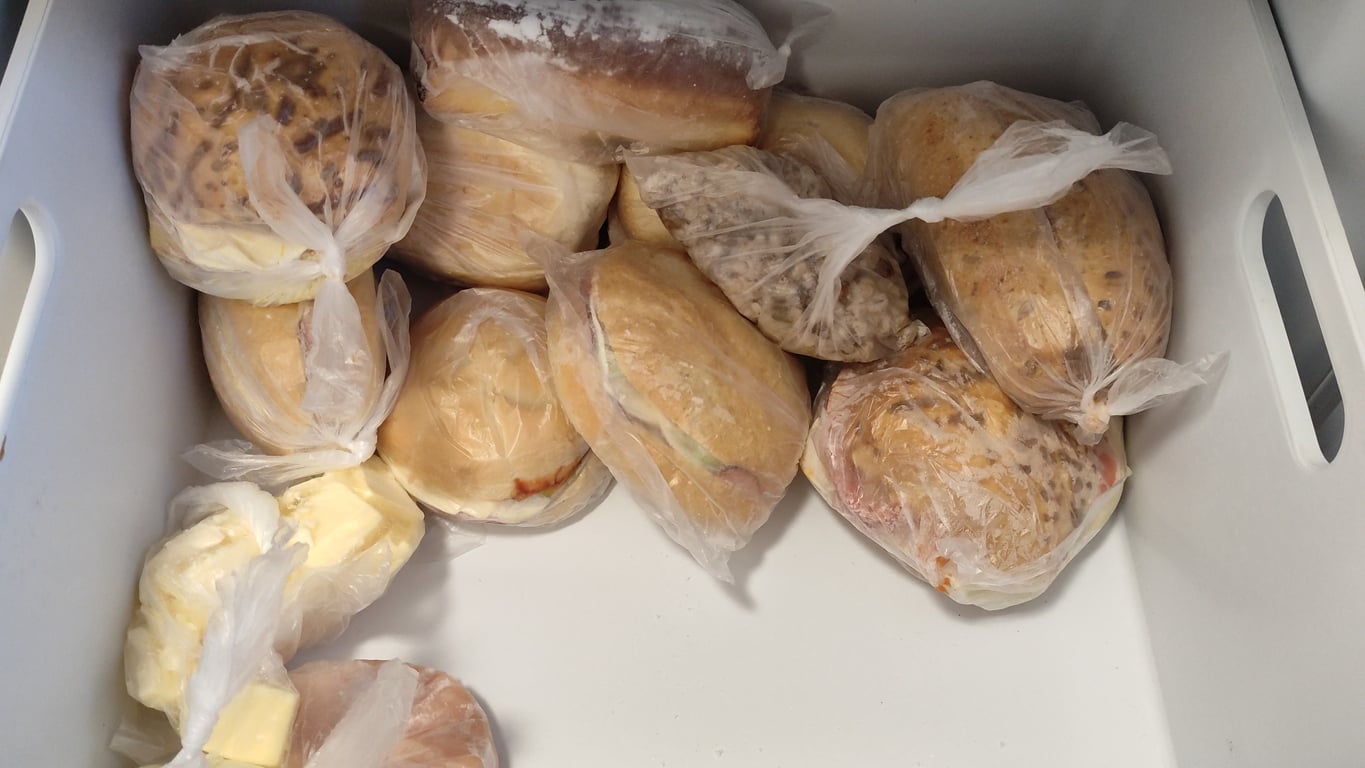 Several sandwiches wrapped in clear plastic bags are placed in a white container. Some sandwiches have visible sesame seed buns, and a few pieces of butter are also included.