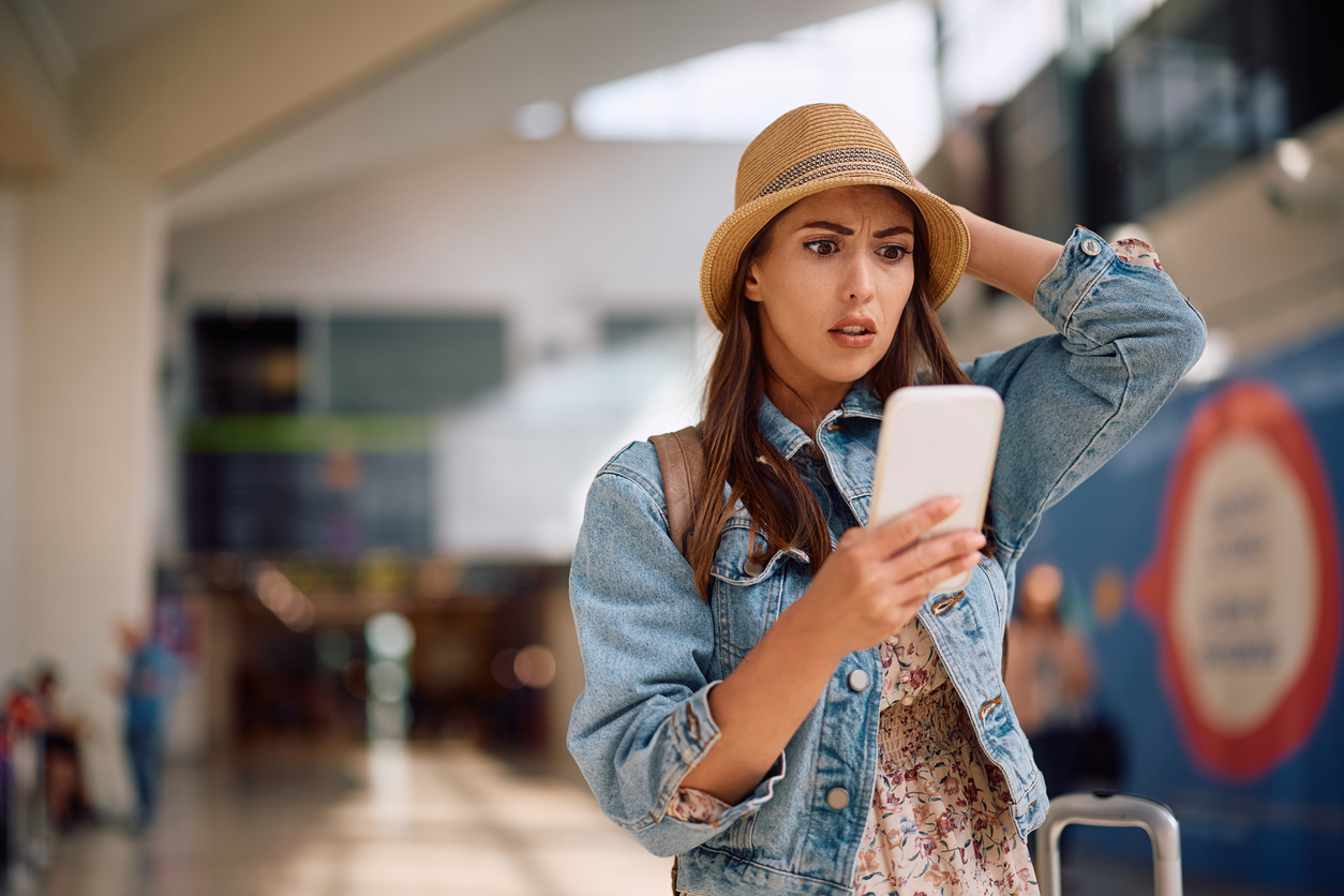A young woman in a denim jacket and hat stands in an airport, looking at her phone with a worried expression while holding her head, suggesting she may have received unexpected or troubling news.