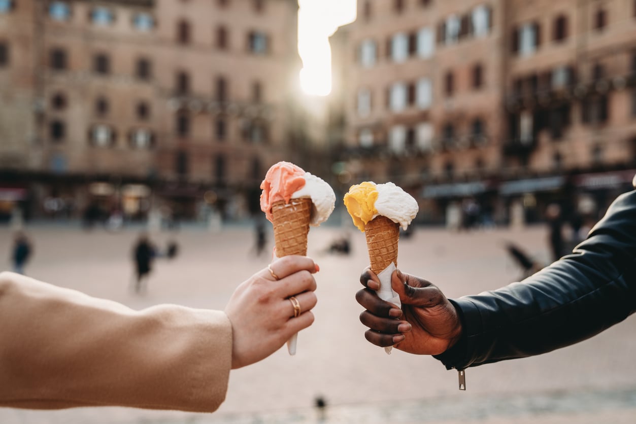 Two people holding ice cream cones with different flavored scoops, raising them towards each other in a toast gesture—celebrating the simple joys that haven’t become products people no longer buy—with a blurred city square in the background.