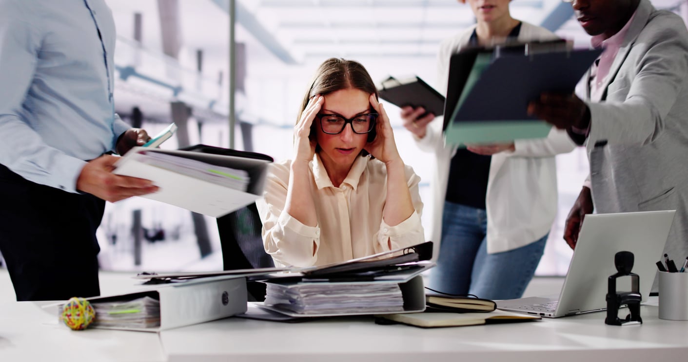 A woman sits at a cluttered desk, holding her head in stress, while three colleagues—reflecting traditional Baby Boomer beliefs about hard work and responsibility—stand around her, handing her documents in a busy office environment.