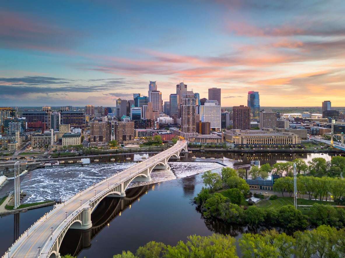 Aerial view of a travel-friendly city skyline at sunset with a bridge crossing over a river, green trees, and buildings reflected in the water under a colorful sky.
