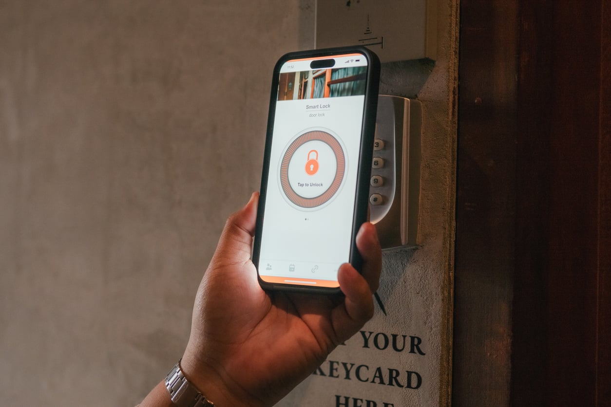 A person holds a smartphone with a smart lock app open, displaying a large lock icon and "Tap to Unlock," in front of an electronic keypad lock, showcasing modern hotel changes in room access technology.