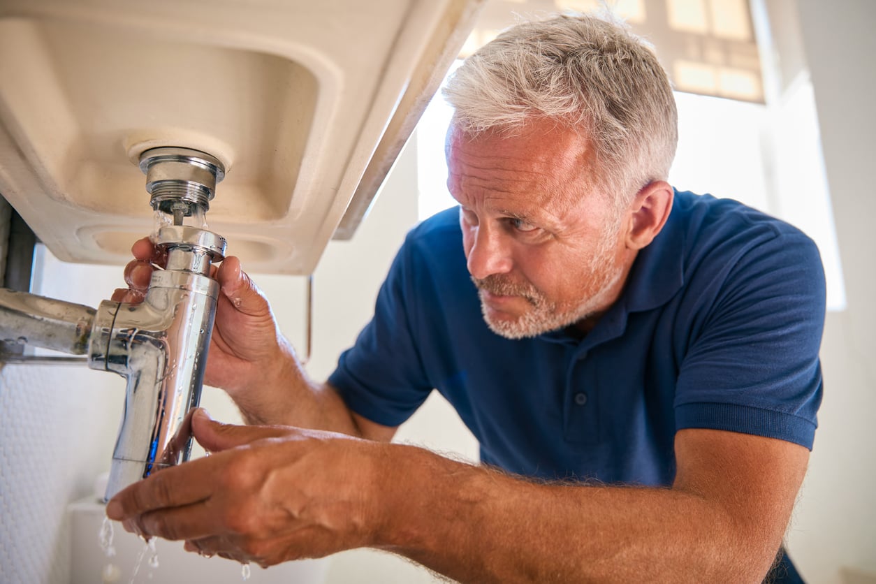 A man in a blue shirt repairs a leaking pipe under a sink, catching dripping water with his hands while inspecting the plumbing closely.