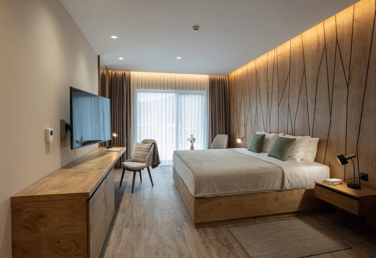 Modern hotel room with a large bed, wooden furniture, a wall-mounted TV, desk, and floor-to-ceiling window with sheer curtains letting in natural light. Warm lighting and neutral tones create a cozy atmosphere—ideal for travelers looking to relax after following essential hotel booking tips to secure the perfect stay.