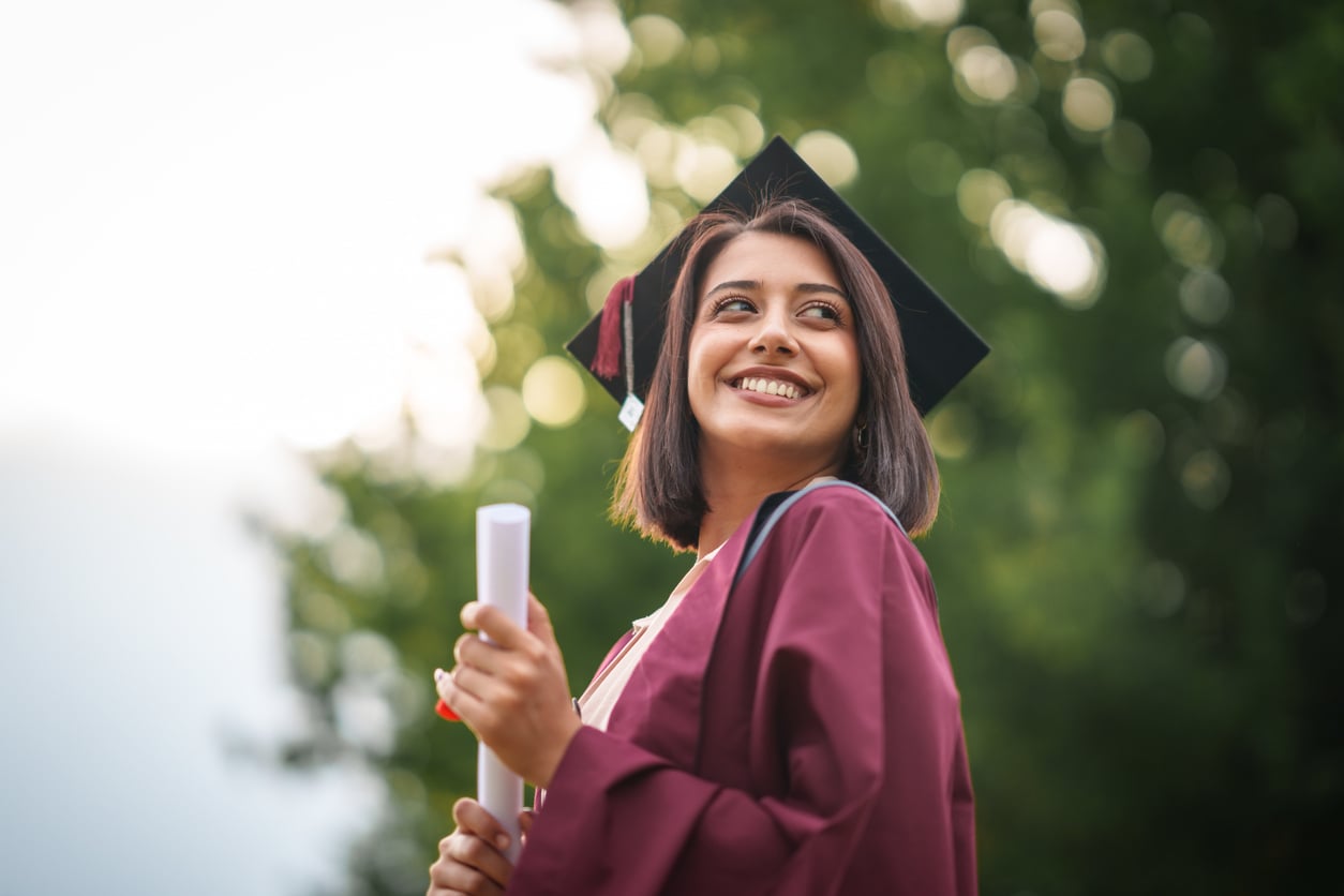 A young woman in a maroon graduation gown and cap smiles while holding a diploma, standing outdoors with green trees in the background—a proud moment that echoes traditional Baby Boomer beliefs in the value of education and hard work.