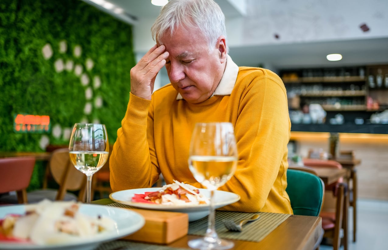 An older man in a yellow sweater sits at a restaurant table, holding his head in his hand and looking stressed, perhaps reflecting on money saving habits, with a plate of food and a glass of white wine in front of him.