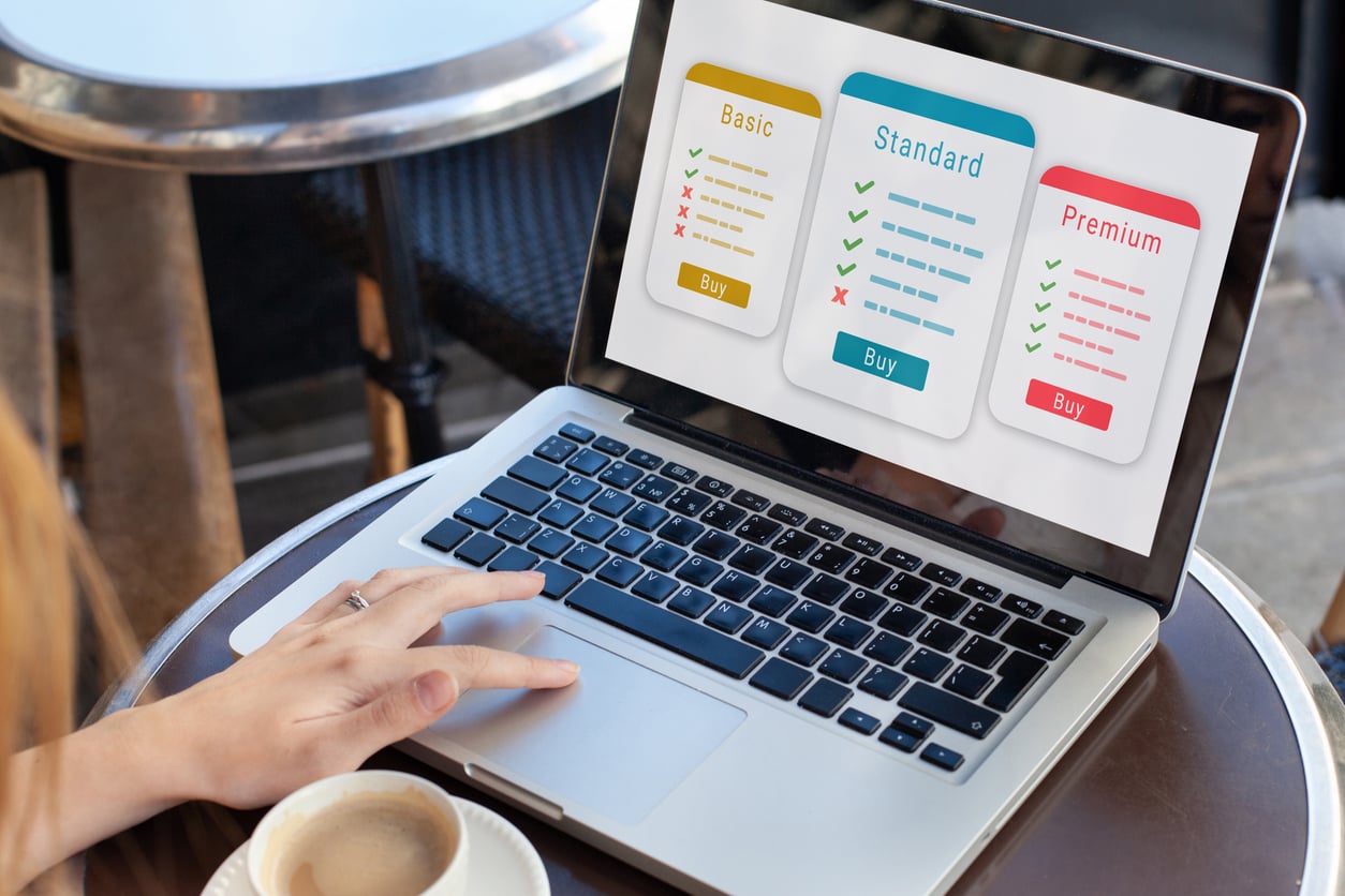 A person using a laptop with a screen displaying a comparison of Basic, Standard, and Premium plans—each plan lists features and has a "Buy" button. A cup of coffee sits next to the laptop as they search for the best movie theater hacks.