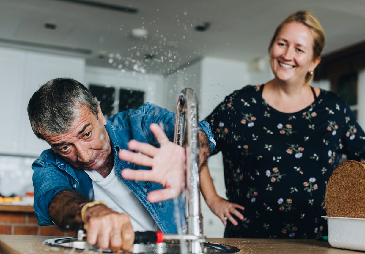 An older man tries to fix a kitchen sink faucet as water sprays everywhere. He raises his hand to block the spray while a woman beside him smiles, amused by the situation.