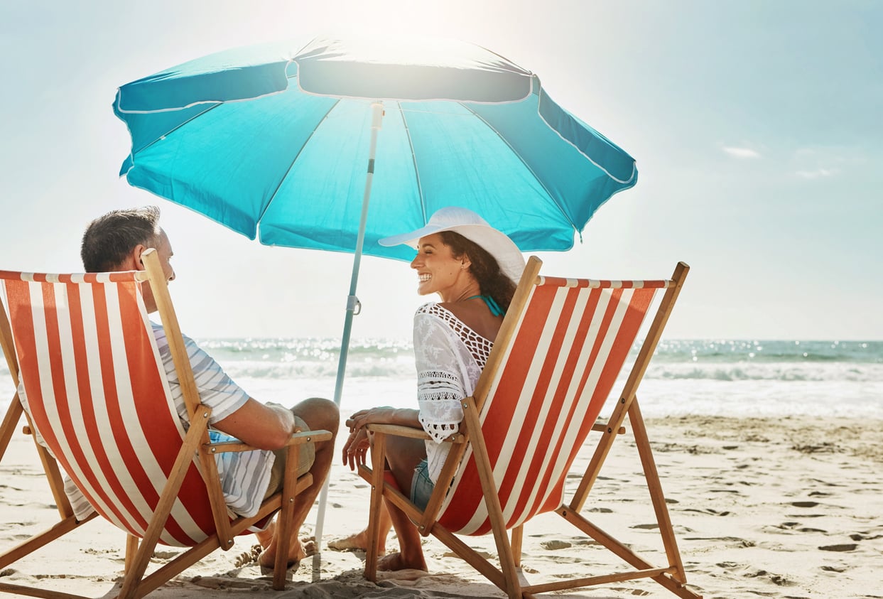 A man and woman sit in striped beach chairs under a blue umbrella, smiling at each other on a sunny day at the beach with sand and ocean in the background, enjoying a carefree moment without worrying about vacation costs.
