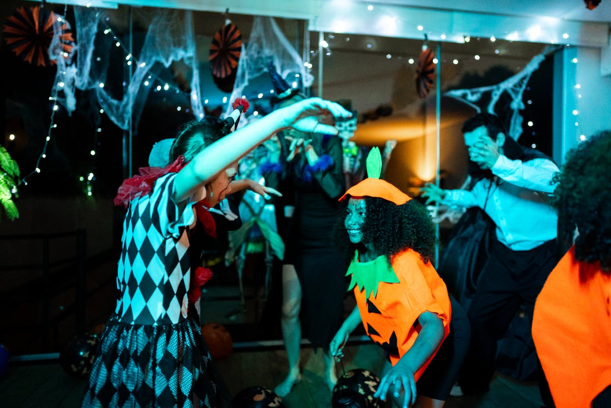Children and adults in Halloween costumes dance and play under string lights, enjoying festive Halloween activities amid decorations like cobwebs and paper swirls in the background.