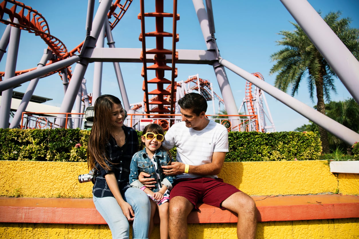 A family of three sits on a bench, smiling and laughing at an amusement park with a roller coaster in the background on a sunny day, enjoying their time together without worrying about vacation costs.