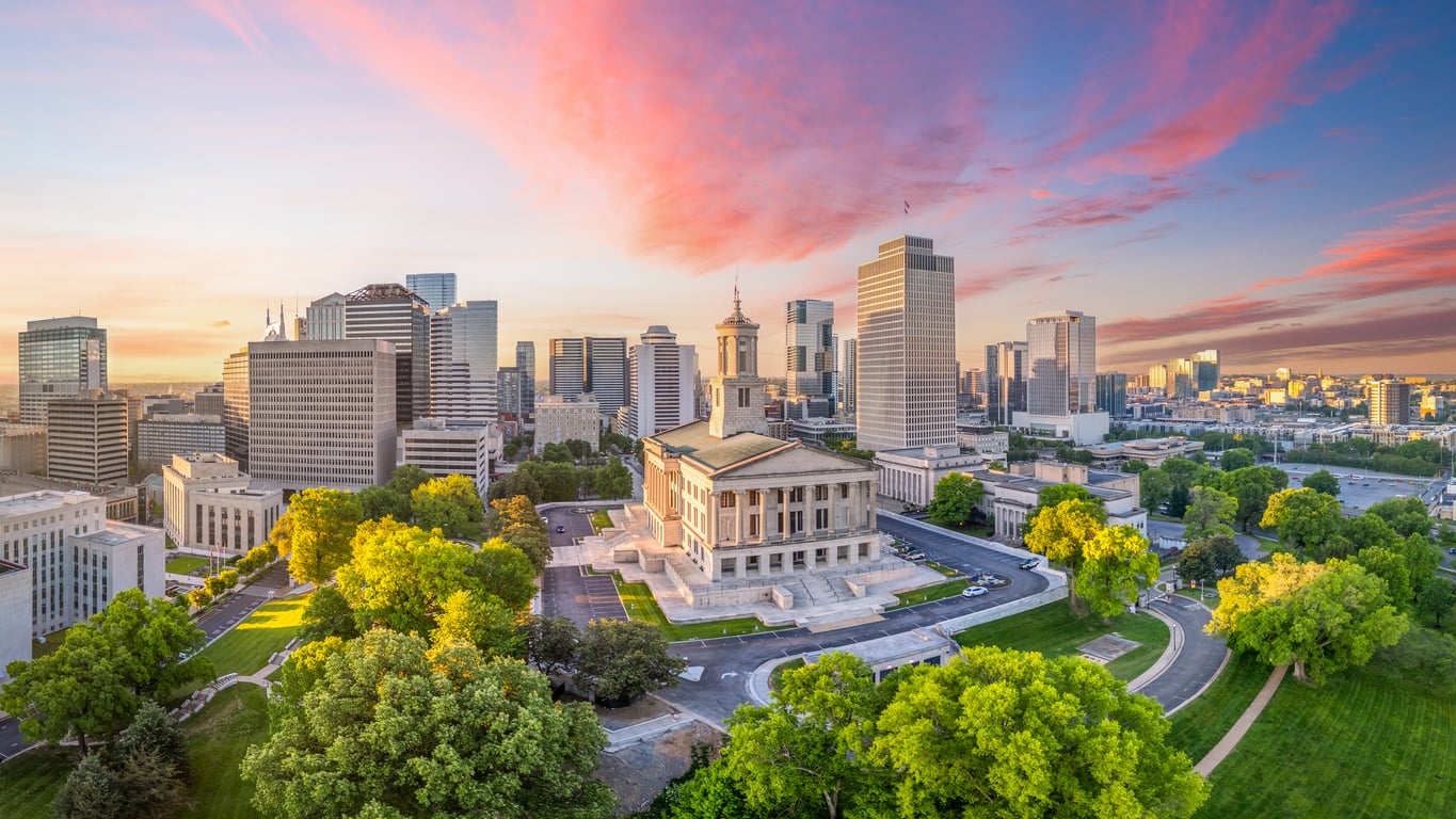 An aerial view of downtown Nashville, Tennessee at sunset, with the Tennessee State Capitol building surrounded by green trees and skyscrapers under a colorful sky.