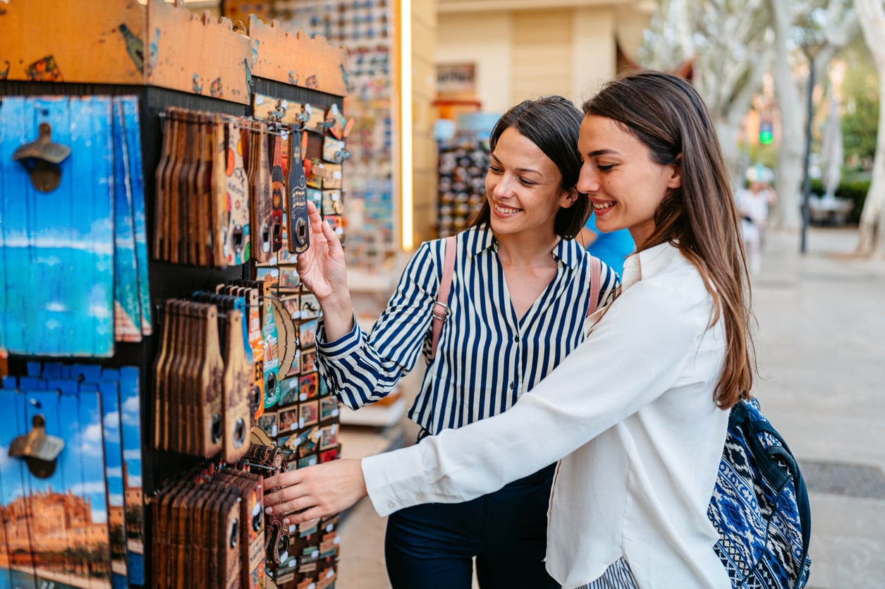 Two women smile while browsing colorful souvenirs at an outdoor market stand on a sunny day, carefully considering vacation costs as they select the perfect keepsake. One woman points at magnets as the other looks on, both appearing happy and engaged. Trees and buildings are visible in the background.