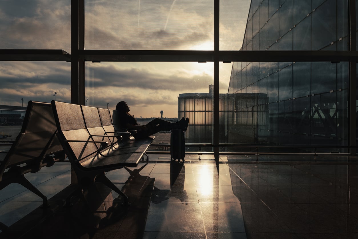 A person sits on airport benches with their feet on a suitcase, silhouetted against large windows at sunset, perhaps taking a quiet moment to reflect before booking a flight.
