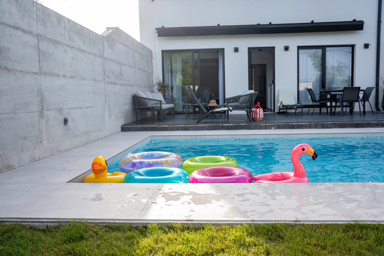 A modern backyard pool with inflatable toys, including a pink flamingo, a yellow duck, and colorful rings—showcasing the benefits of renting a home that comes with great outdoor amenities. The pool sits beside a patio in front of a white house.