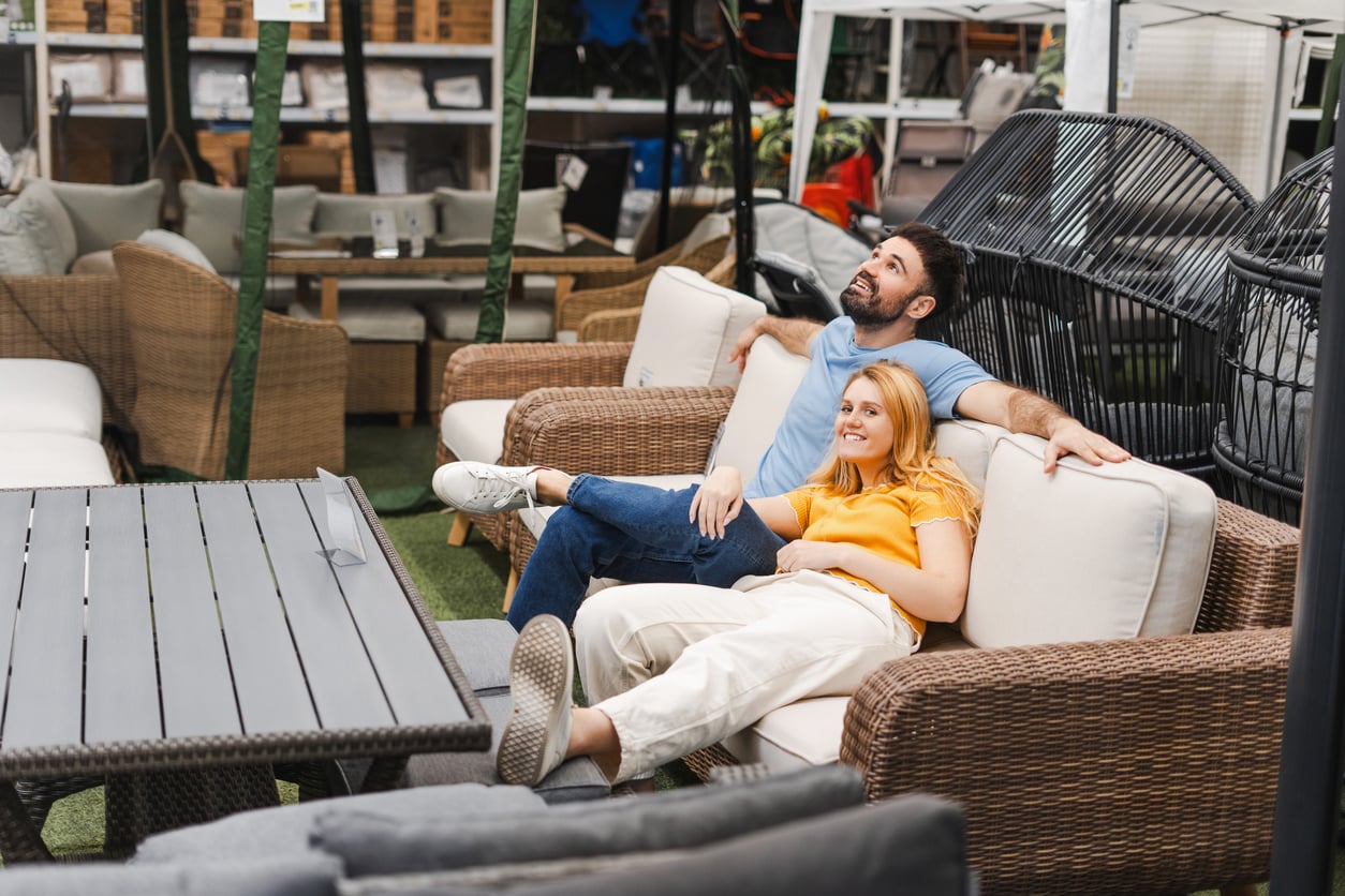 A man and woman relax together on a wicker outdoor sofa in a furniture store, smiling and looking up. Surrounded by other patio furniture sets—some reminiscent of products people no longer buy—they enjoy the comfort and style of their choice.