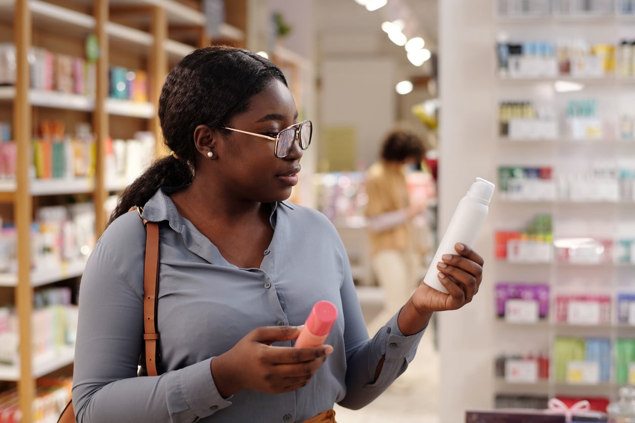 A woman wearing glasses examines two bottles while shopping in a brightly lit store aisle filled with various products on shelves, reflecting how Women Are Changing Their Spending Behaviors.