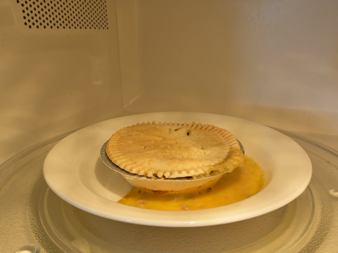 Frozen food products. A pot pie on a white plate sits inside a microwave, with filling spilling out onto the plate from under the crust.