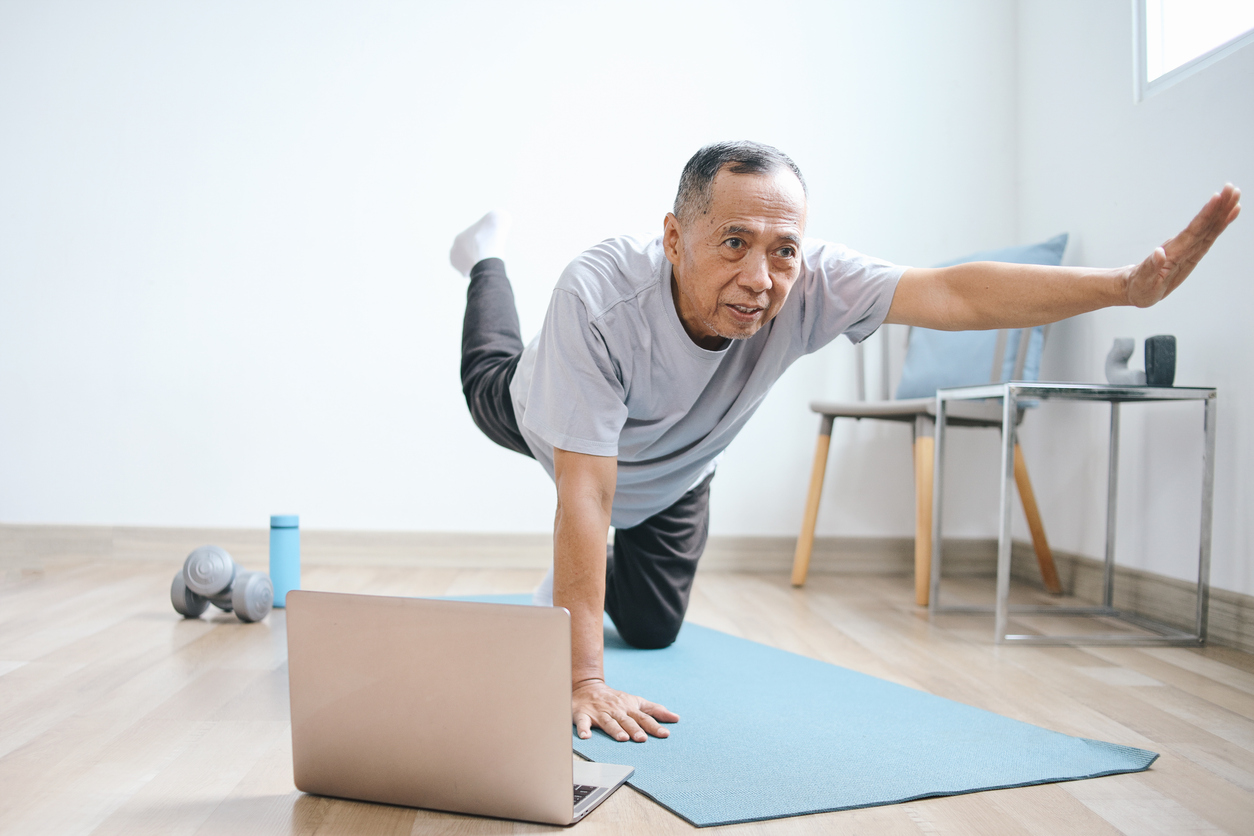 An older man exercises on a yoga mat at home, balancing on one knee and hand with his opposite arm and leg extended. By working out at home, he saves money while using his laptop, water bottle, and dumbbells in the sunlit, bright room.